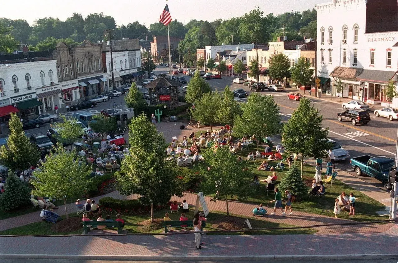 Town square park with many people, trees, benches, and surrounding buildings under an American flag.