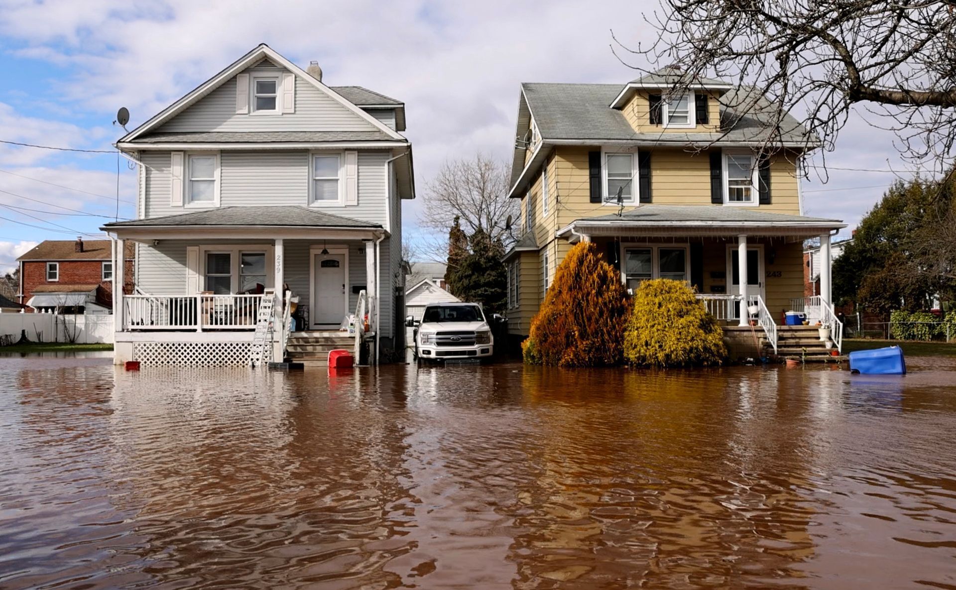 Flooded street with two houses, brown water covering the road and parked vehicles partially submerged.