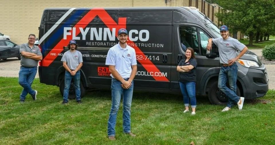 Five people stand in front of a black FLYNNCO service van on a grassy street.