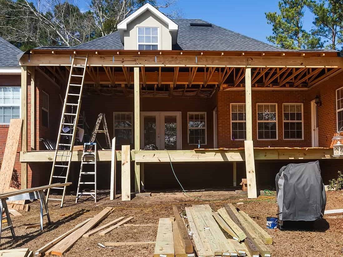 House porch under construction with ladders, exposed framing, and stacked lumber in front