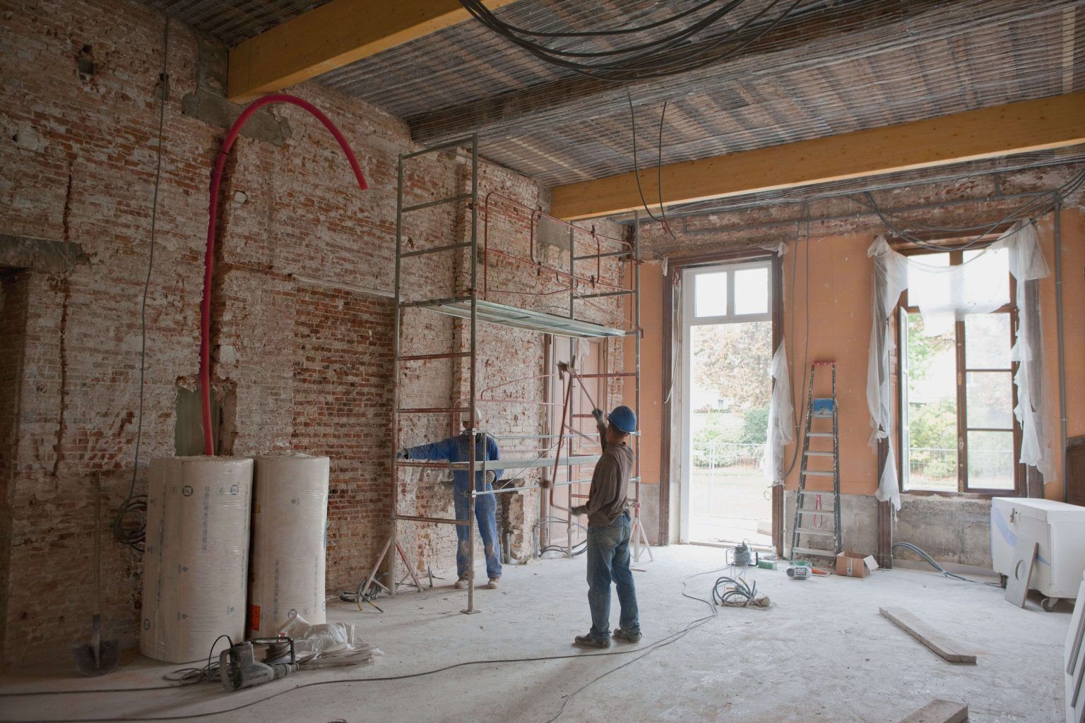 Worker in an unfinished brick room with exposed pipes, large windows, and construction materials.