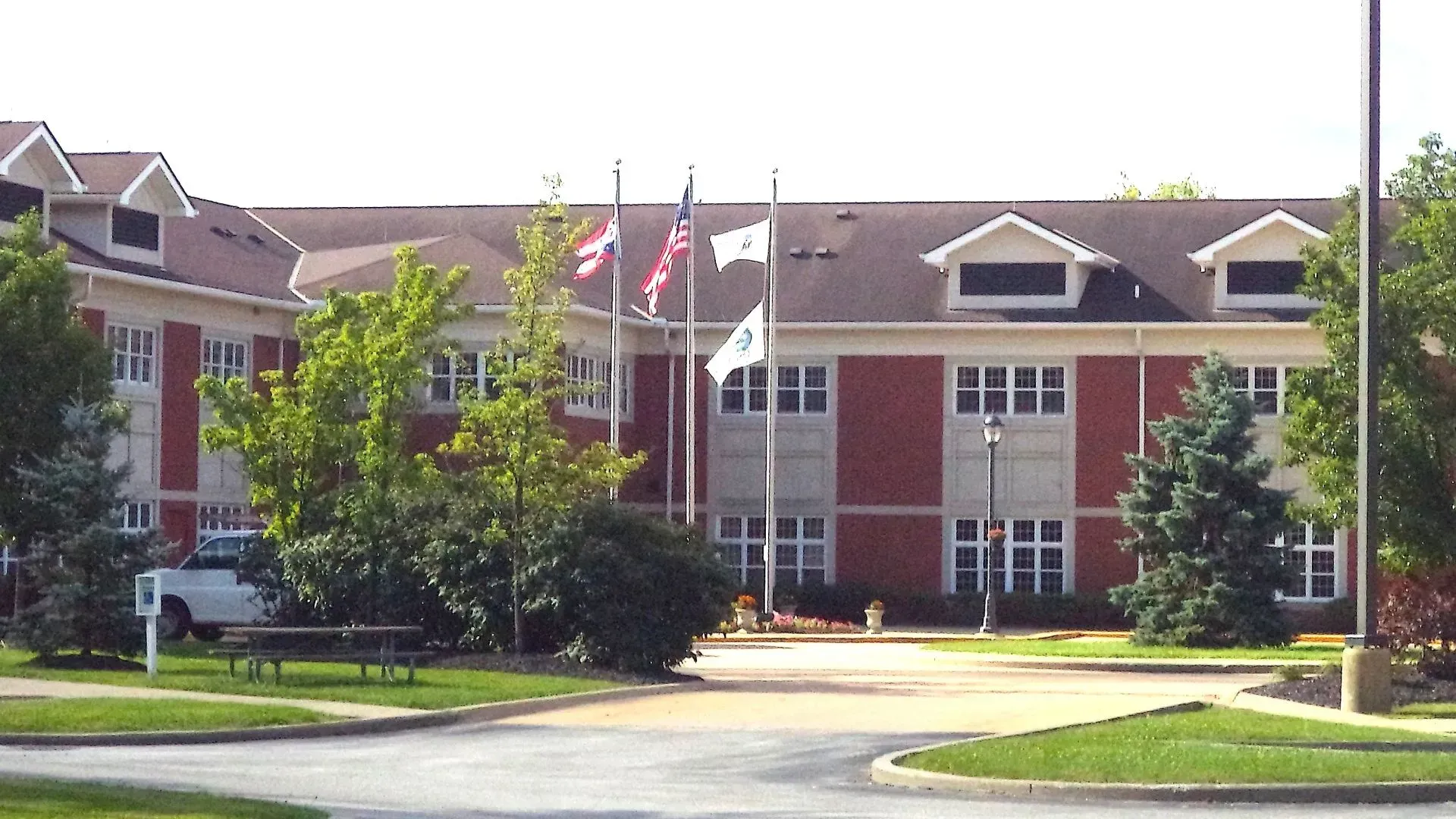 Brick apartment buildings with flagpoles and a driveway surrounded by green trees and lawns