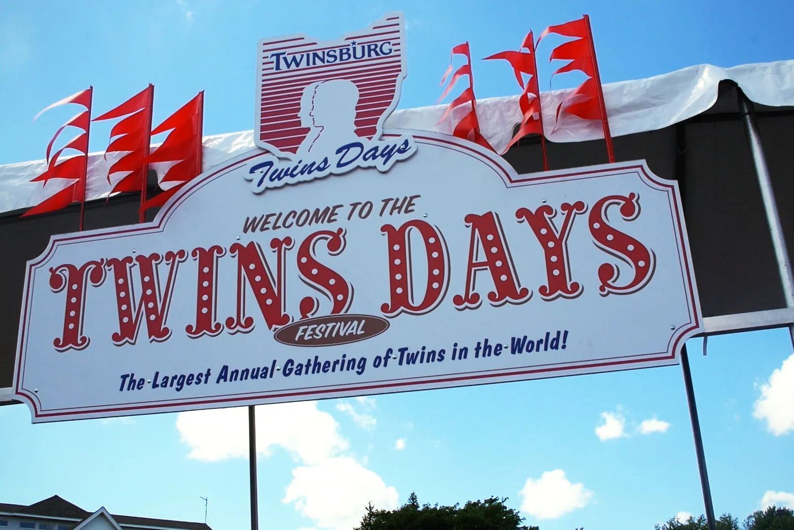 Banner reading “Twins Days” with red flags against a blue sky, welcoming attendees to the festival