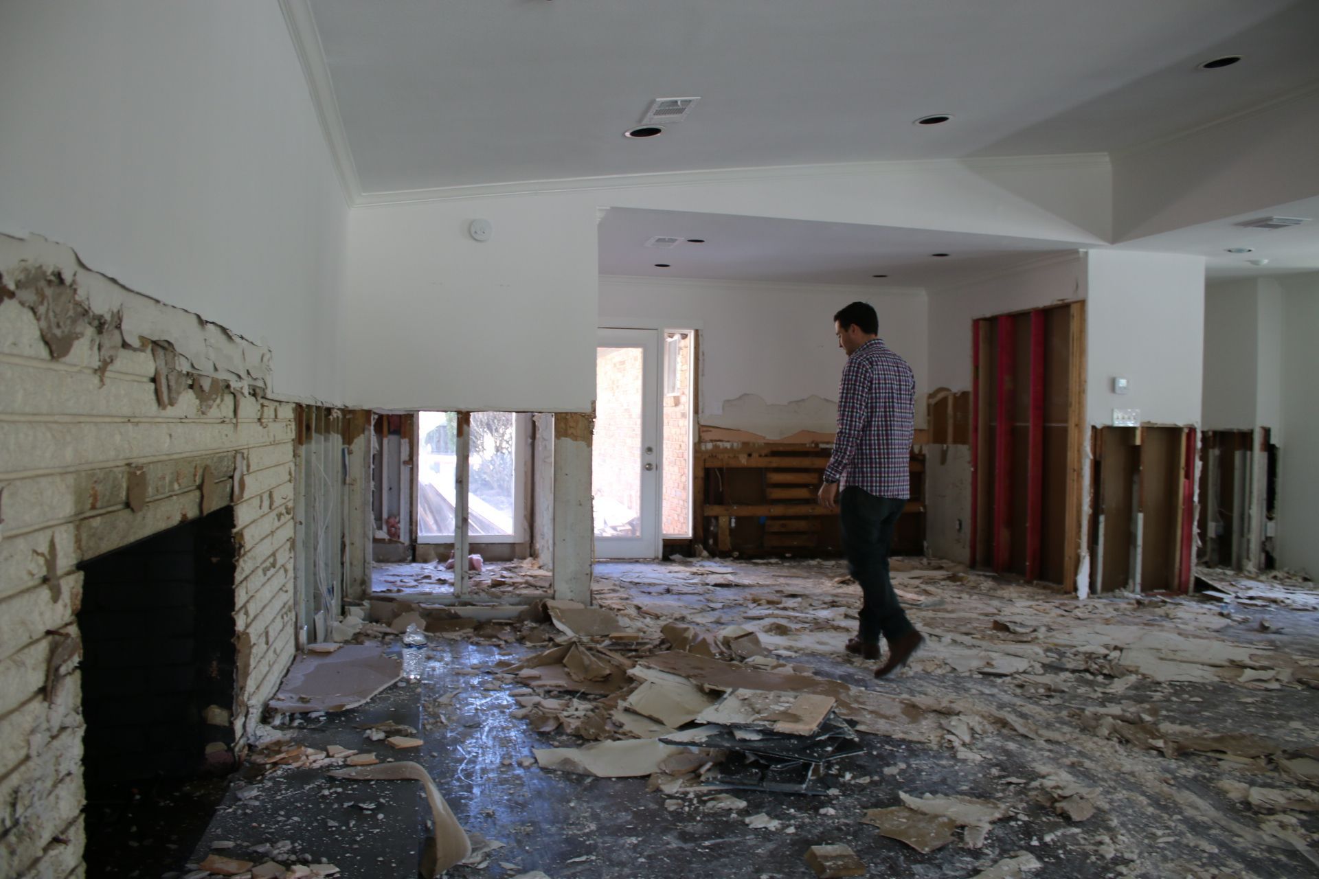 Damaged living room with debris-covered floor and a person standing near a fireplace and exposed walls