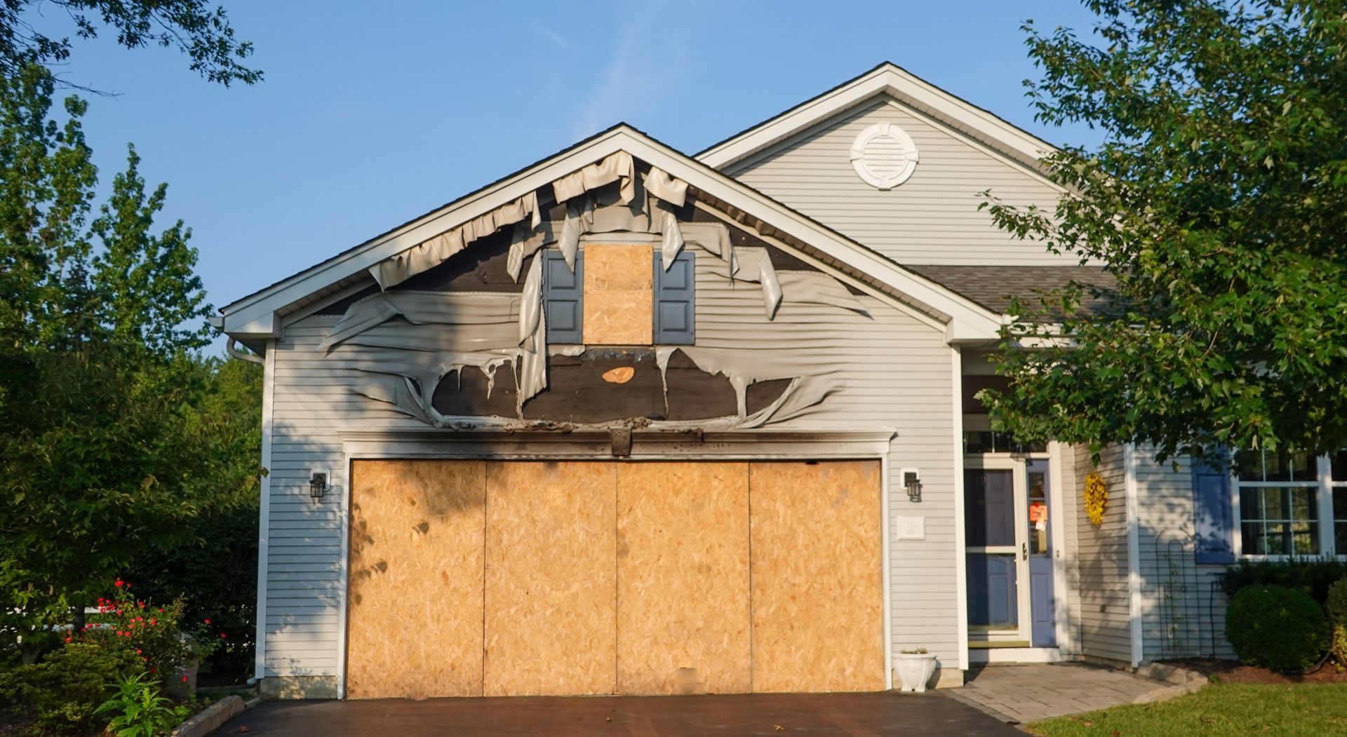House with boarded-up garage door and damaged upper facade in a suburban neighborhood