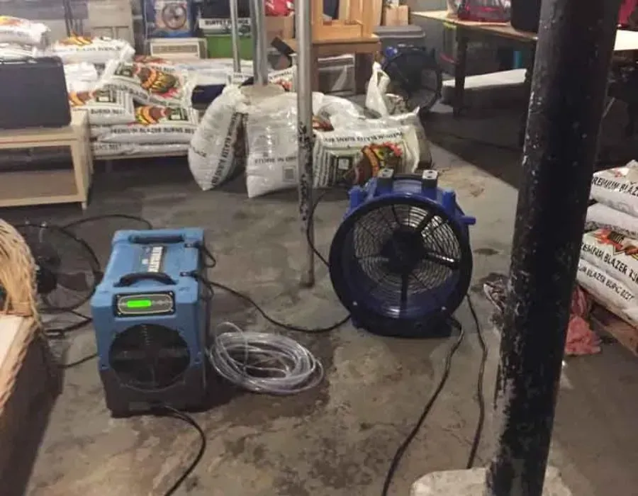 Blue portable fan and black blower on a concrete floor in a cluttered indoor workspace.