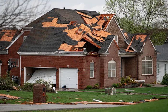 House roof severely damaged by storm, with torn shingles and debris scattered in front yard.