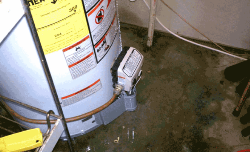 Water heater in a basement next to a floor drain and small pump, with water pooling on the floor.