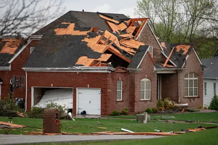 Damaged brick house with roof torn off, debris scattered on lawn after storm