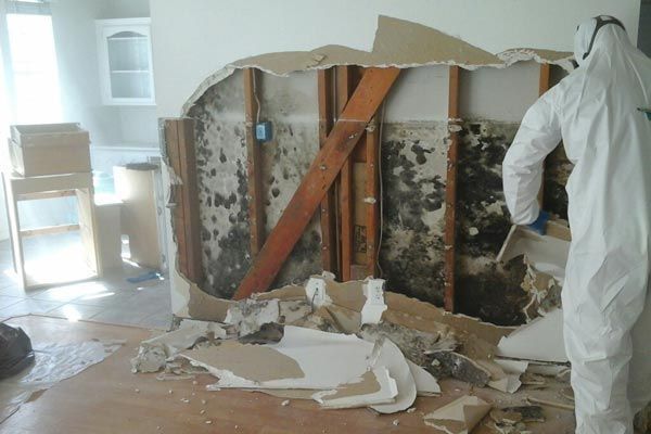 Worker in white protective suit inspects wall demolition with exposed studs and debris in a room