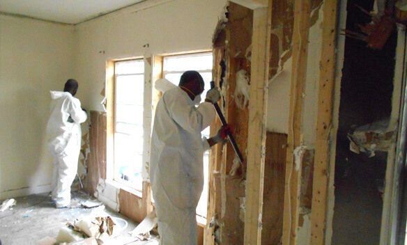 Workers in white protective suits demolish an interior wall in a dusty room.