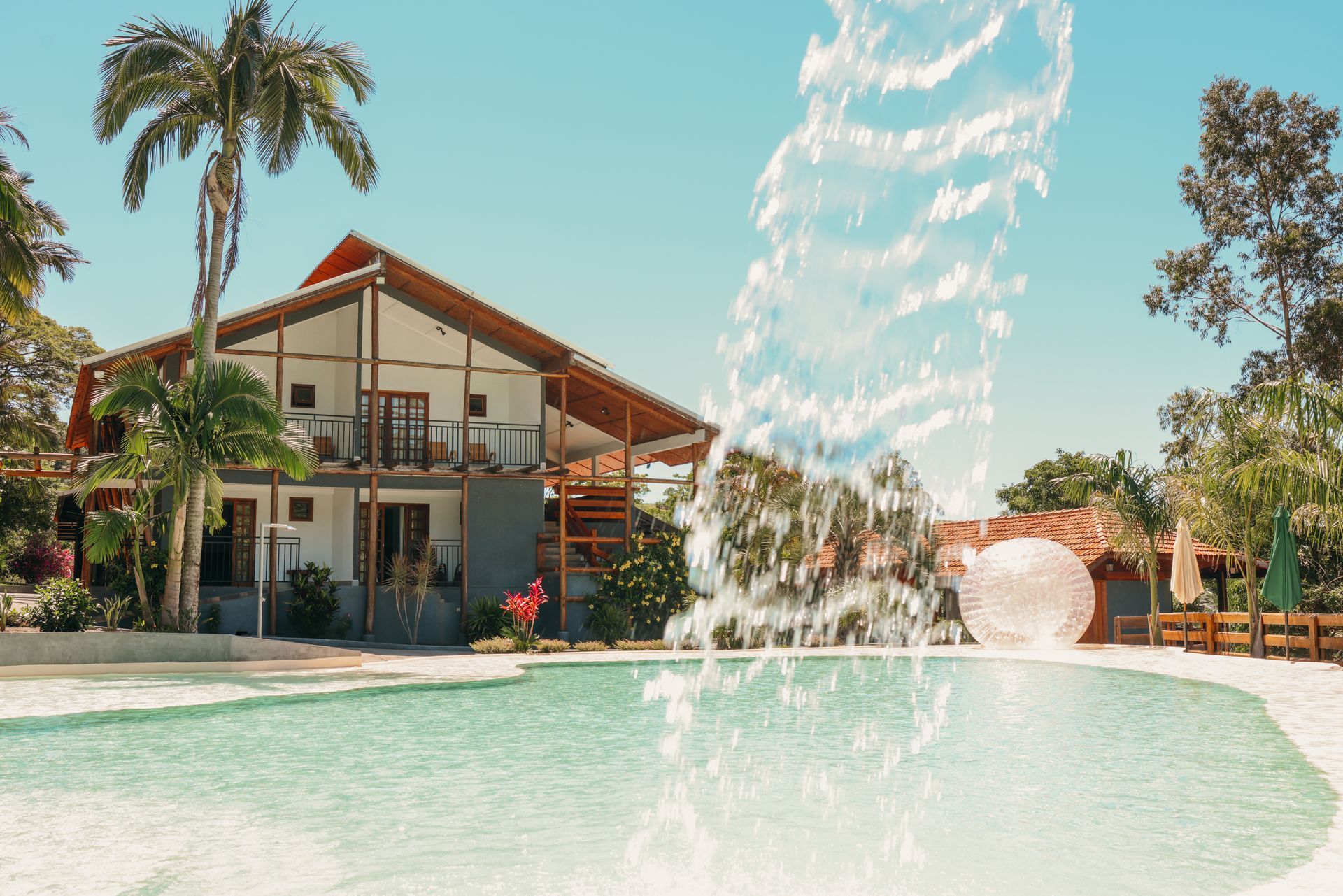 Um resort com piscina, cachoeira e um prédio de dois andares sob um céu azul e ensolarado.