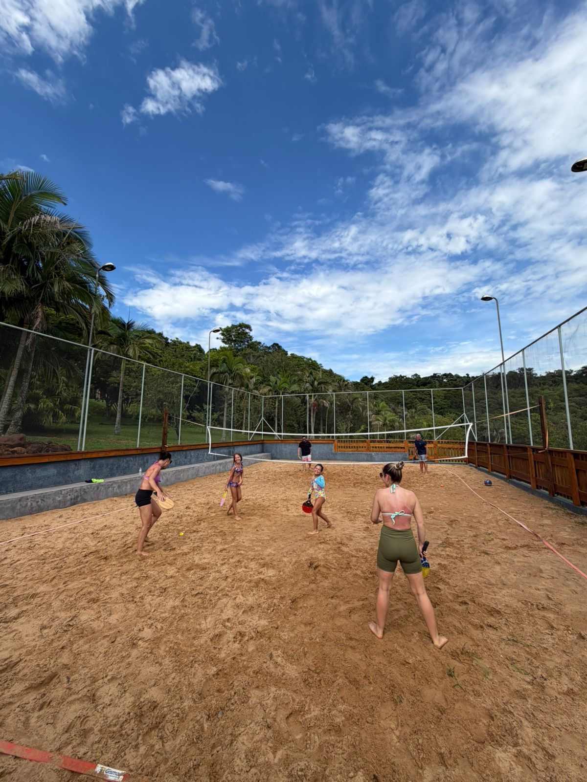 Pessoas jogando tênis de praia em uma quadra de areia sob um céu azul, cercadas por árvores e uma cerca.
