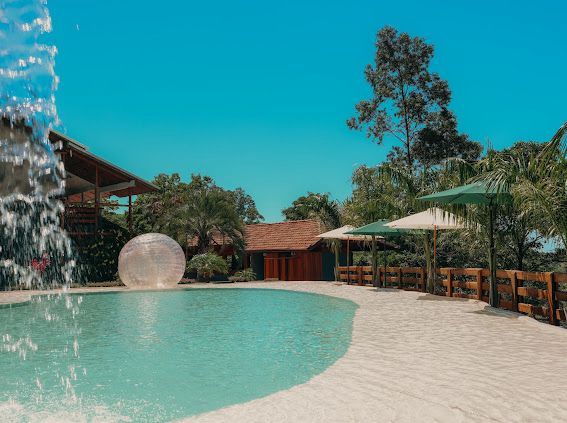 Piscina com borda de areia branca, água azul, cascata e pequenas construções sob um céu azul brilhante.