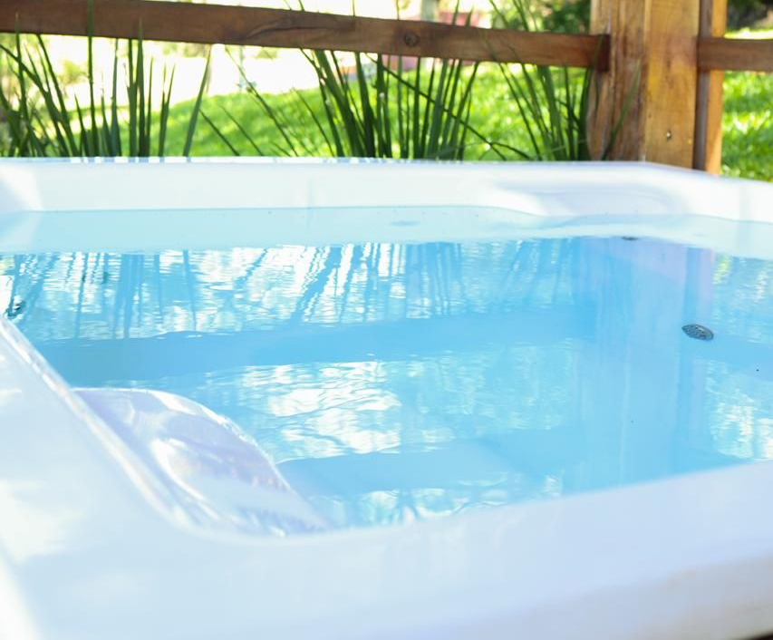 Banheira de hidromassagem cheia de água azul, com uma cerca de madeira e grama verde visíveis ao fundo.