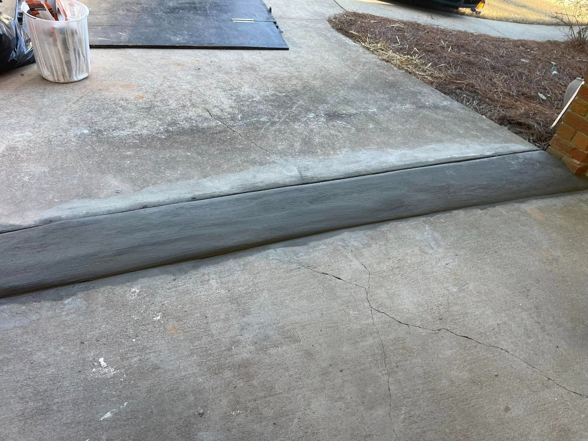 Concrete sidewalk ramp at a curb beside a driveway, with a slight slope and rough-edged repair.
