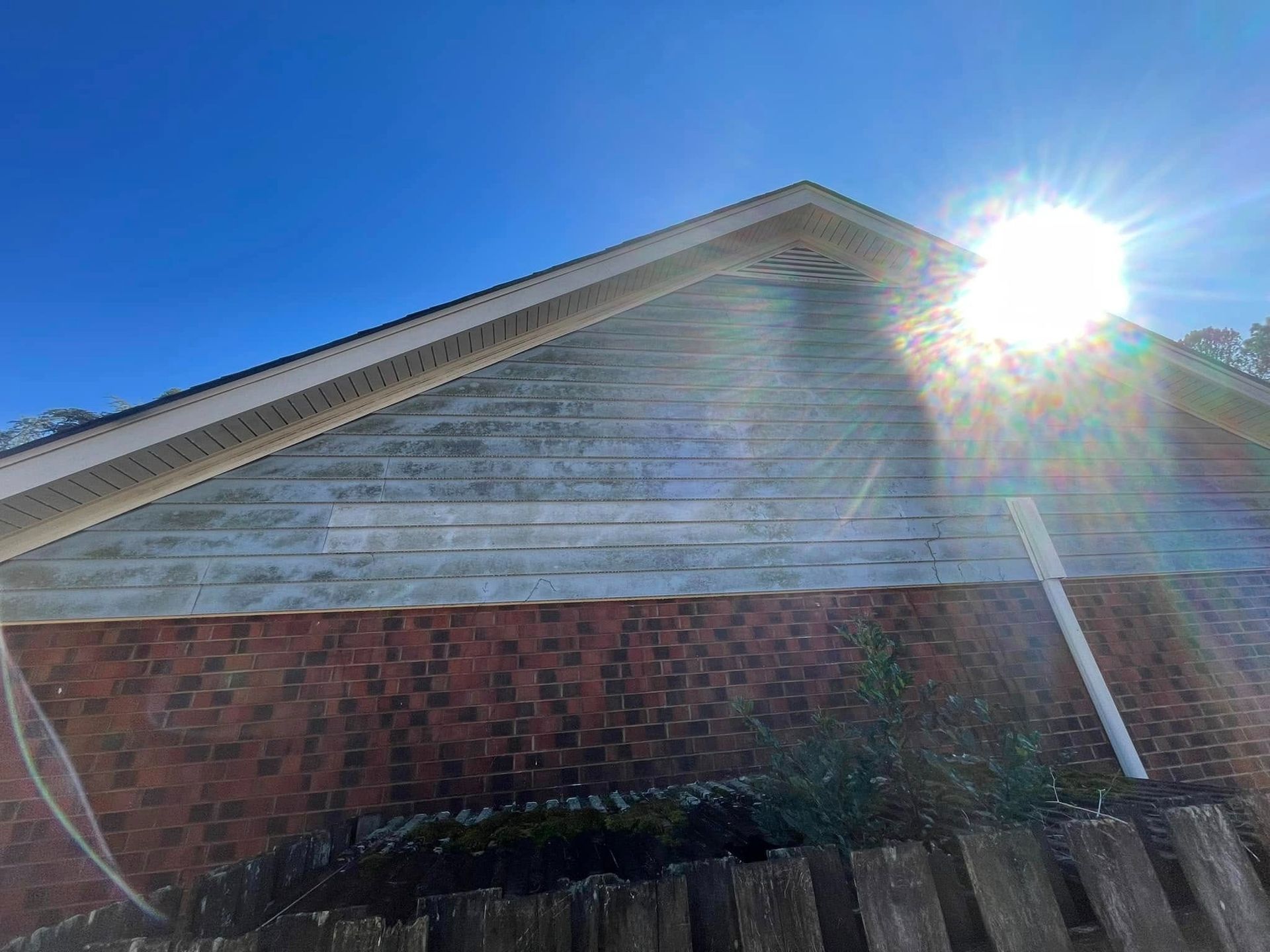 Brick house gable with weathered white siding, bright sun flare in the upper right, and blue sky above