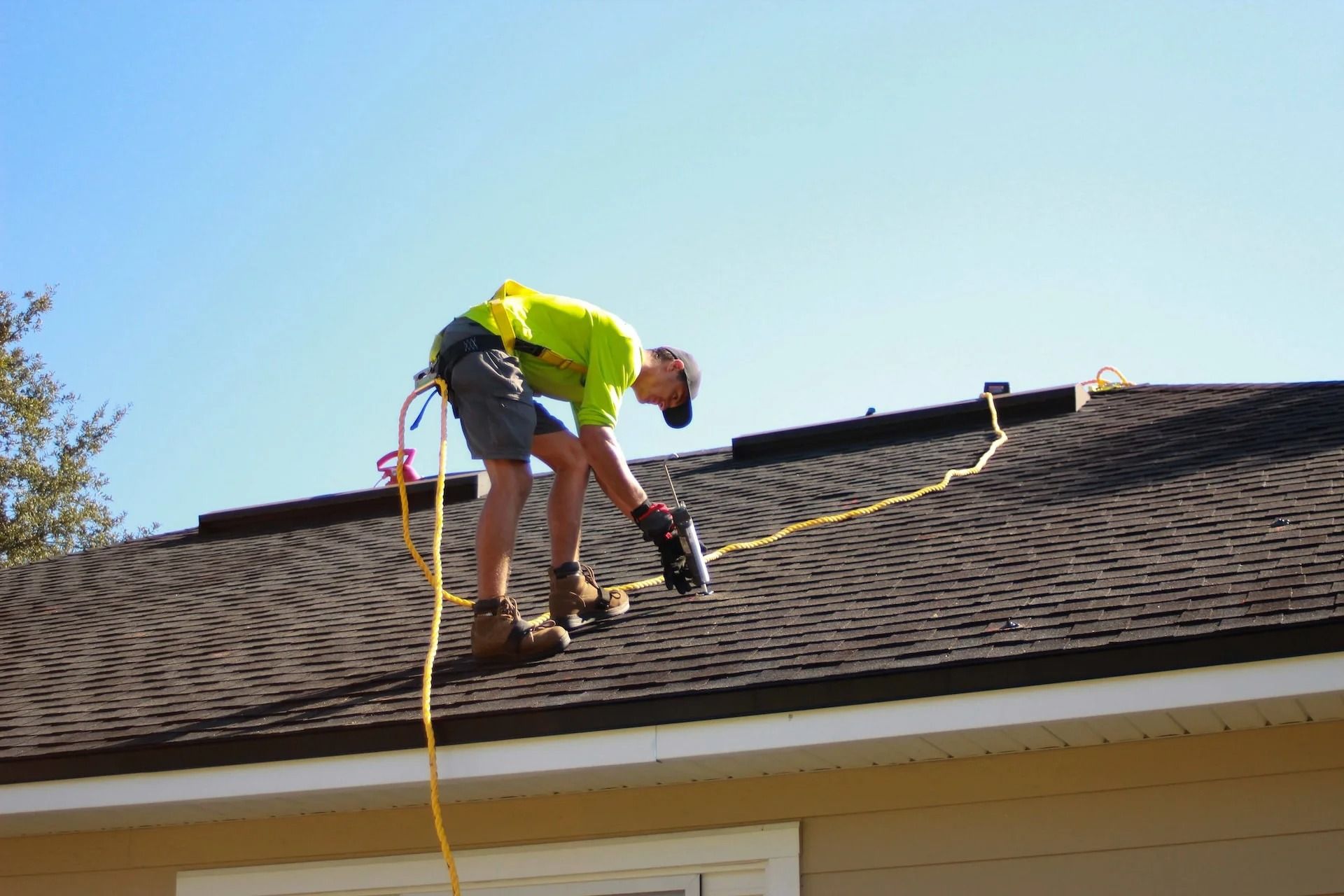 Worker using a power tool on a shingled roof, with safety harness and yellow hose.