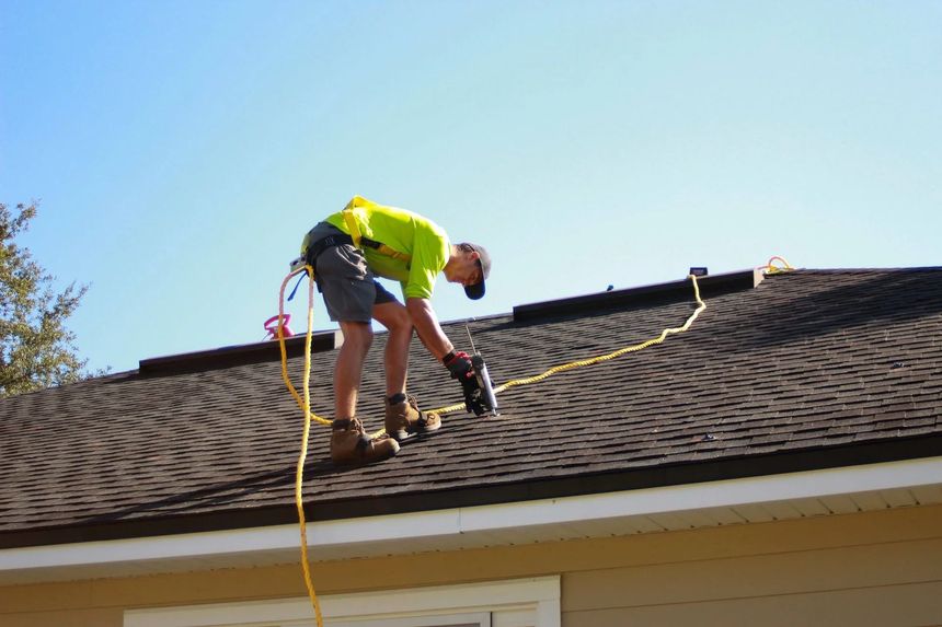 Worker on a shingled roof using a nail gun with safety harness and extension cord
