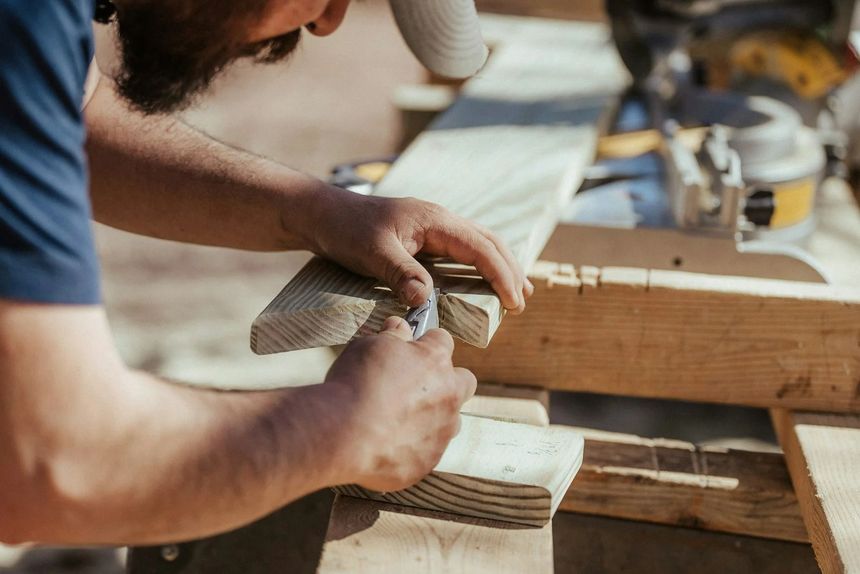 Person hand-sanding a wooden plank in a workshop with a saw in the background