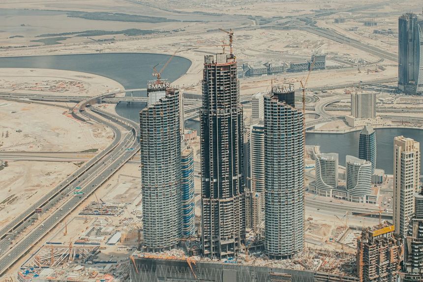 High-rise towers under construction in a sandy desert cityscape, with roads and a waterfront nearby