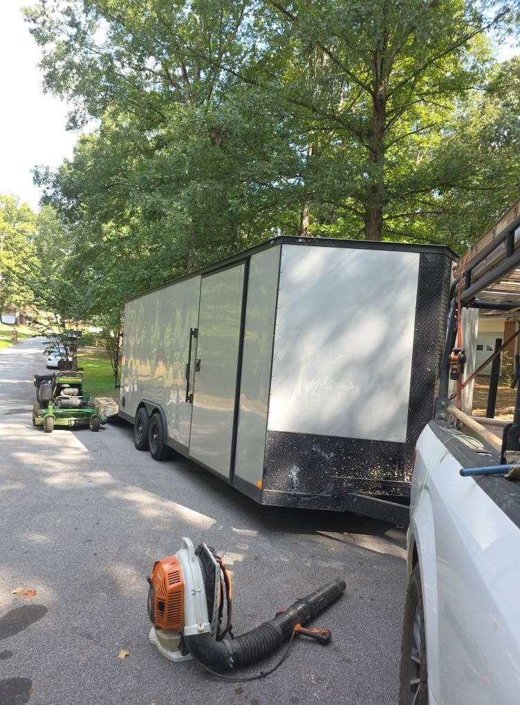 White box trailer parked on a tree-lined street, with a leaf blower and person’s legs visible in the foreground