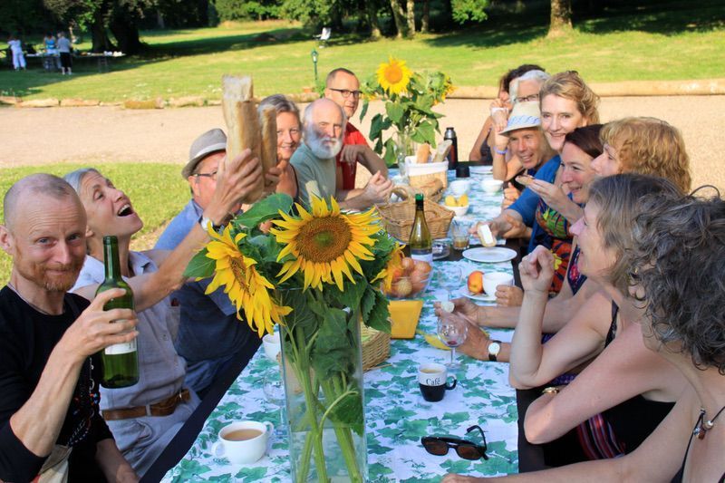 Een groep uiteenlopende mensen viert feest aan een lange tafel met zonnebloemen en eten, terwijl ze lachen en het glas heffen.