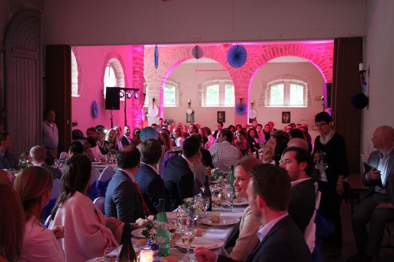 Een grote feestzaal vol mensen, zittend aan tafels, verlicht door roze en witte lampen.
