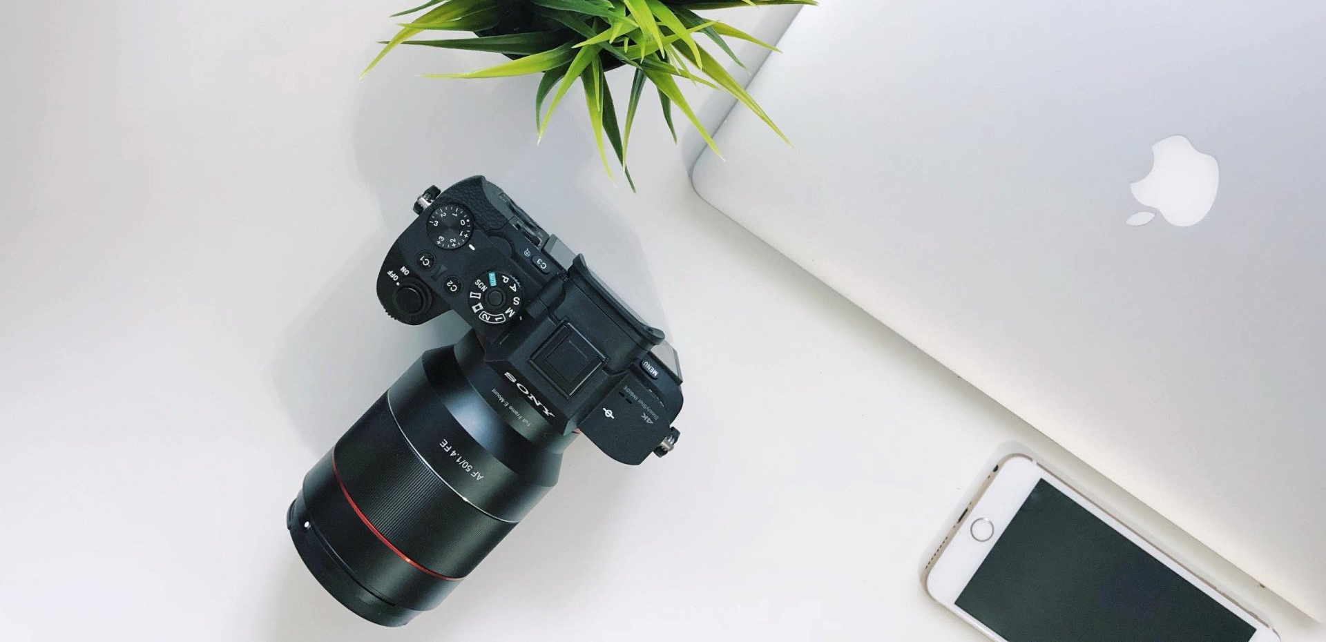 A plant, a DSLR Camera, a macbook laptop, and an iPhone on a white table