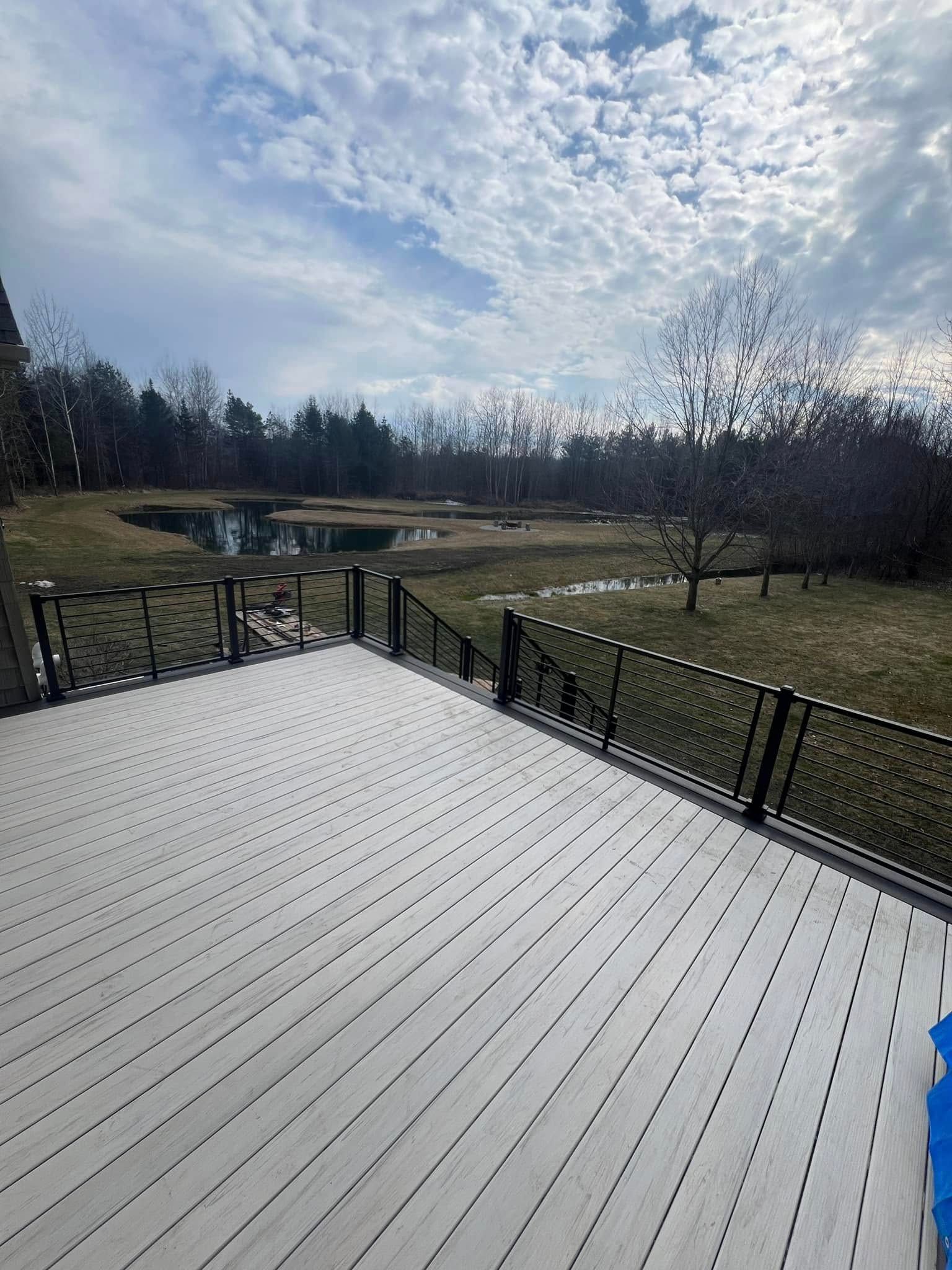 Wooden deck overlooking a grassy yard, trees, and a cloudy sky near a small bridge.