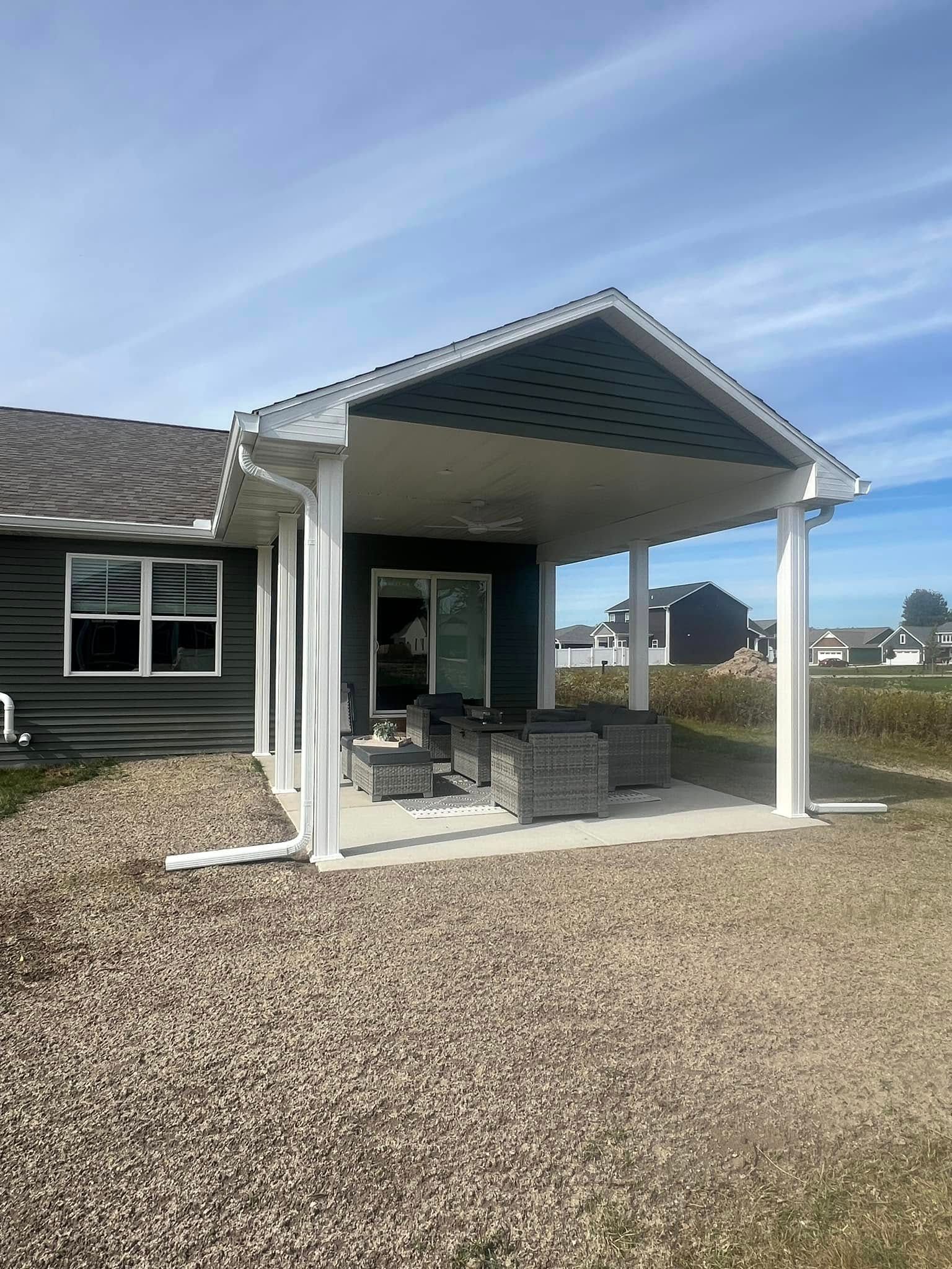 Covered patio with white posts, outdoor seating, and gravel yard beside a house under a blue sky
