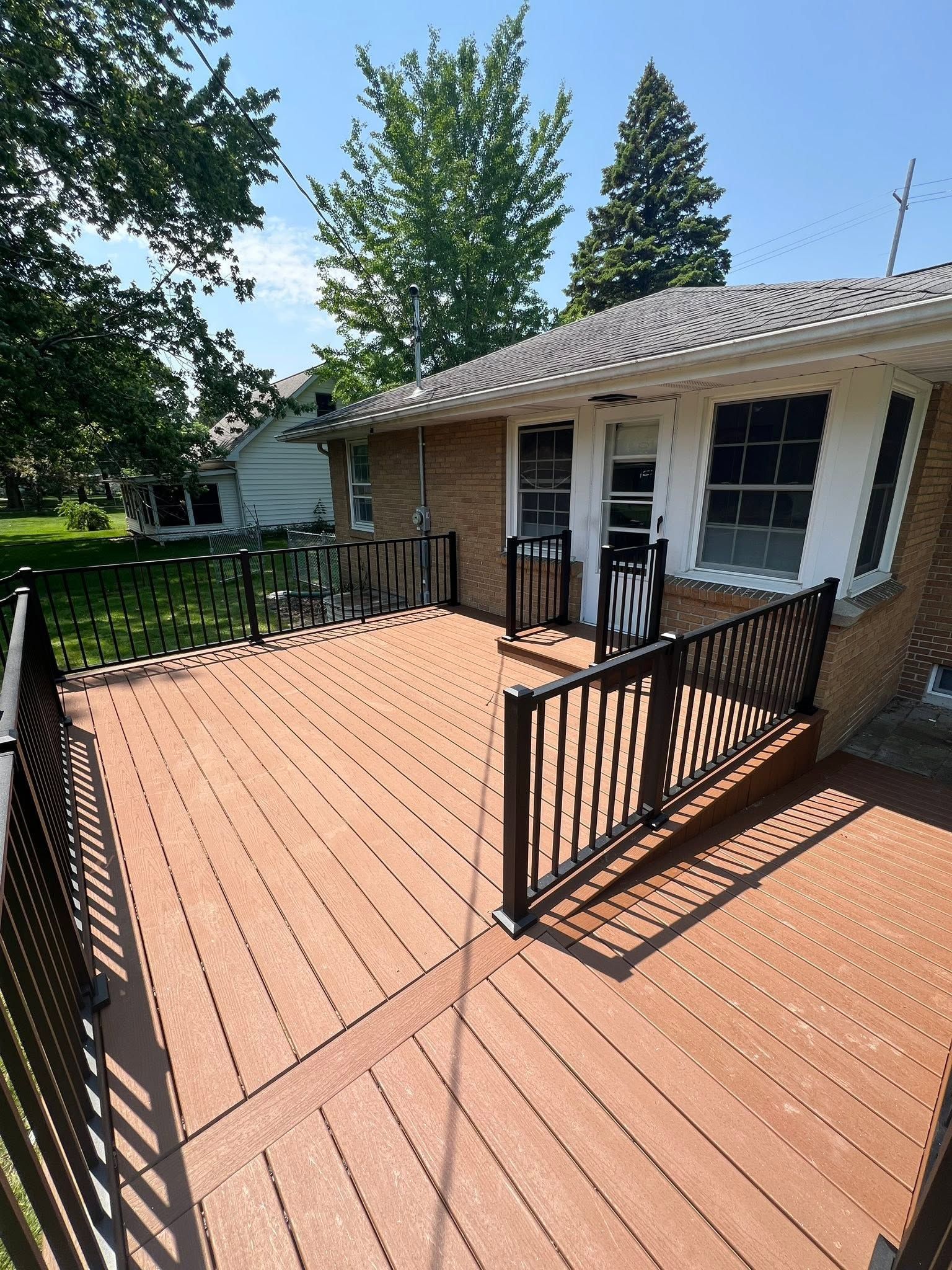 Wooden deck with black railing beside a beige house and white shutters, surrounded by trees and yard.
