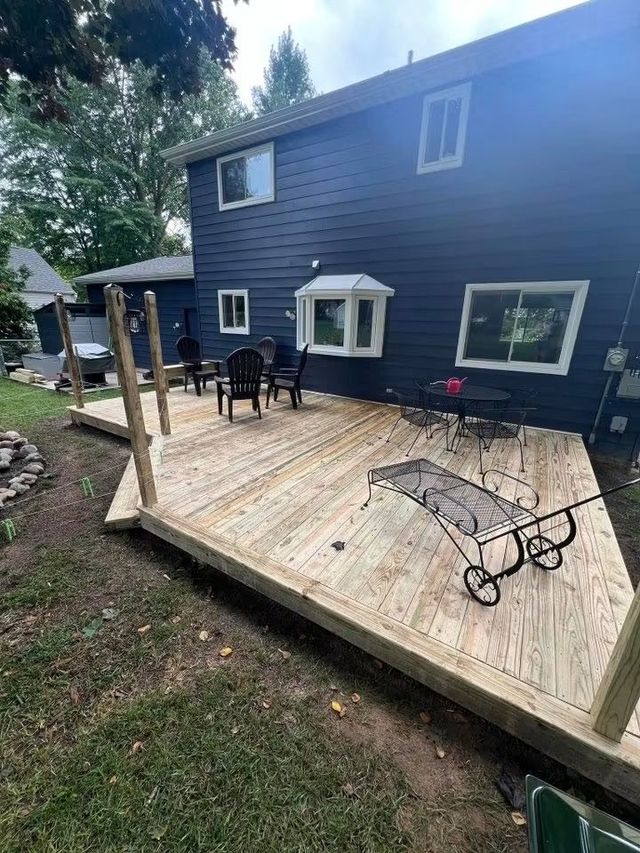 Wooden backyard deck beside a dark blue house, with patio chairs and a small table.