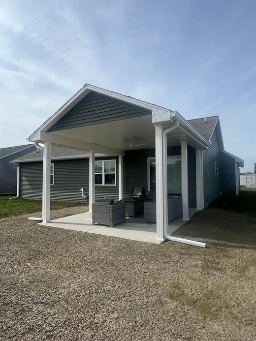 Small white porch pavilion with a peaked roof on a gravel lot under a blue sky