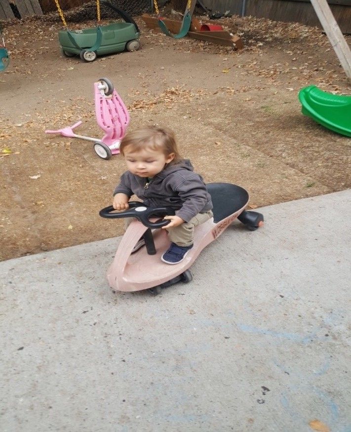 A little boy is riding a pink toy car on a sidewalk.