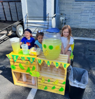 Three kids smiling at a lemonade stand. Sunny day; yellow and green decor, and clear pitcher of lemonade.