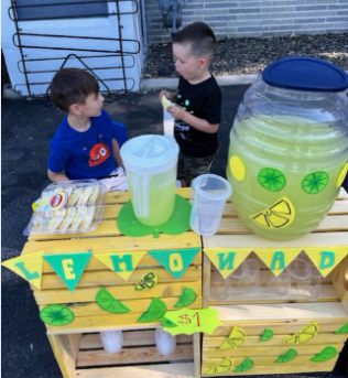 Two young boys running a lemonade stand: yellow stand, green decorations, $1 sign.