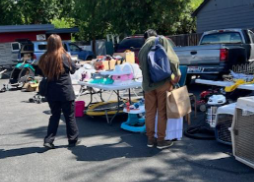 People shopping at an outdoor yard sale with tables of items and parked vehicles on a sunny day.