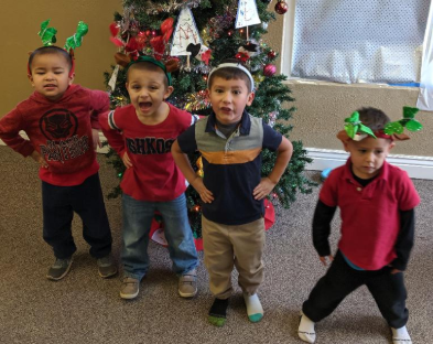 Four young boys wearing reindeer antlers are posing in front of a christmas tree