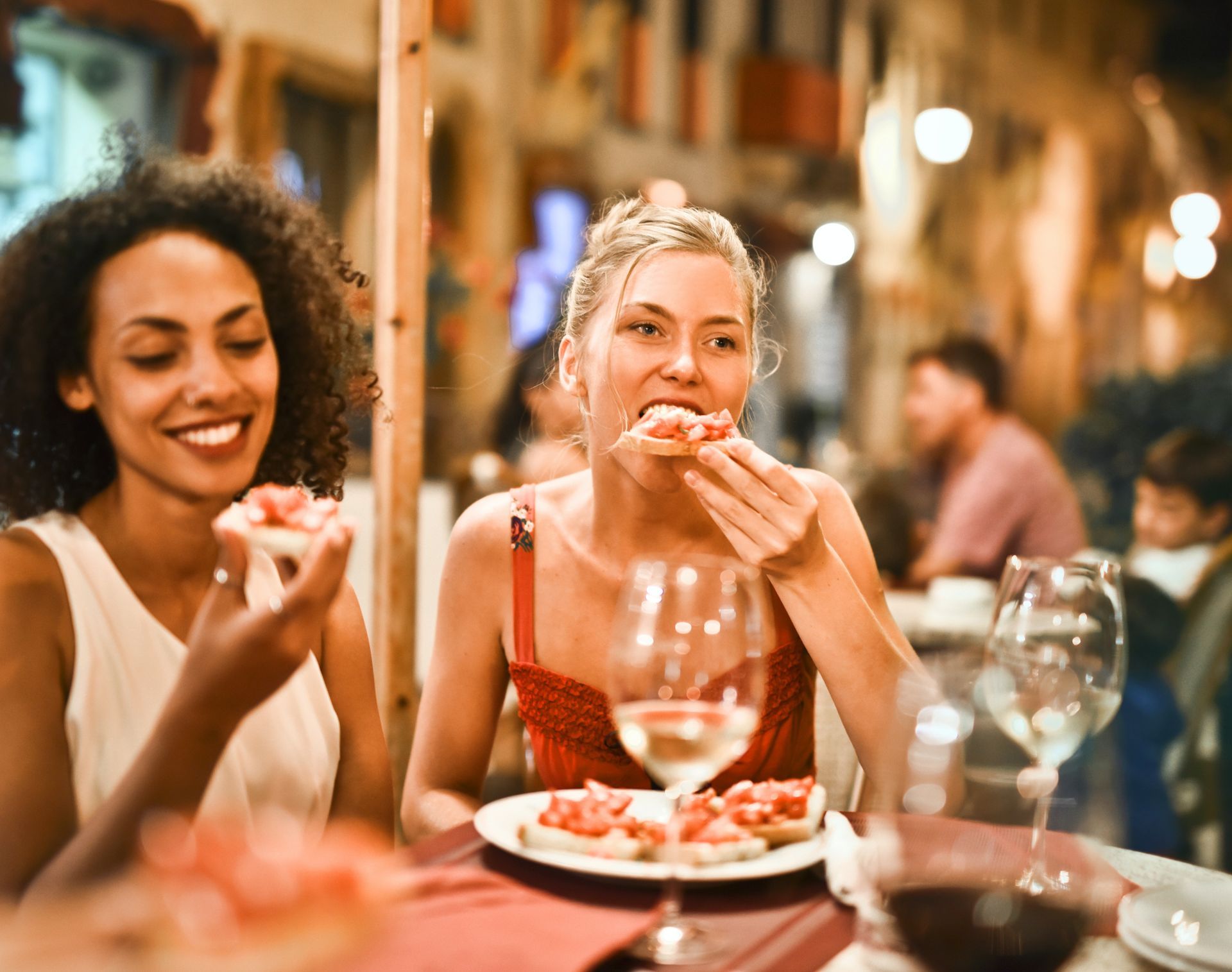 Two women eating bruschetta at a restaurant. One smiles, the other bites hers.