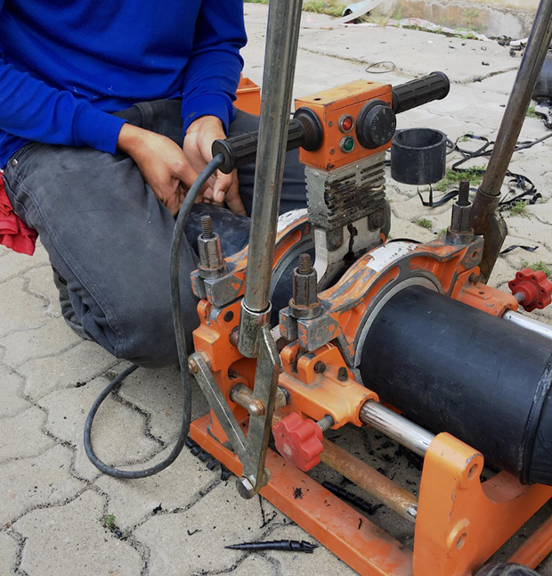 Man using a pipe fusion machine to join black plastic pipes on a paved surface. Orange machine, blue shirt.