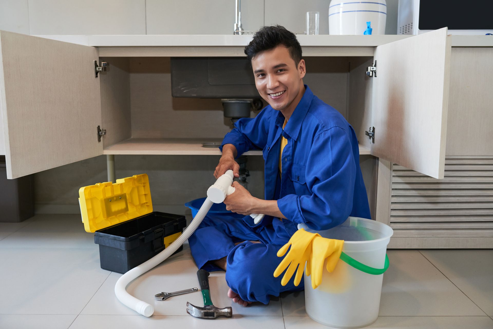 Plumber kneeling under a kitchen sink, smiling, holding a drain snake, with tools and a bucket nearby.