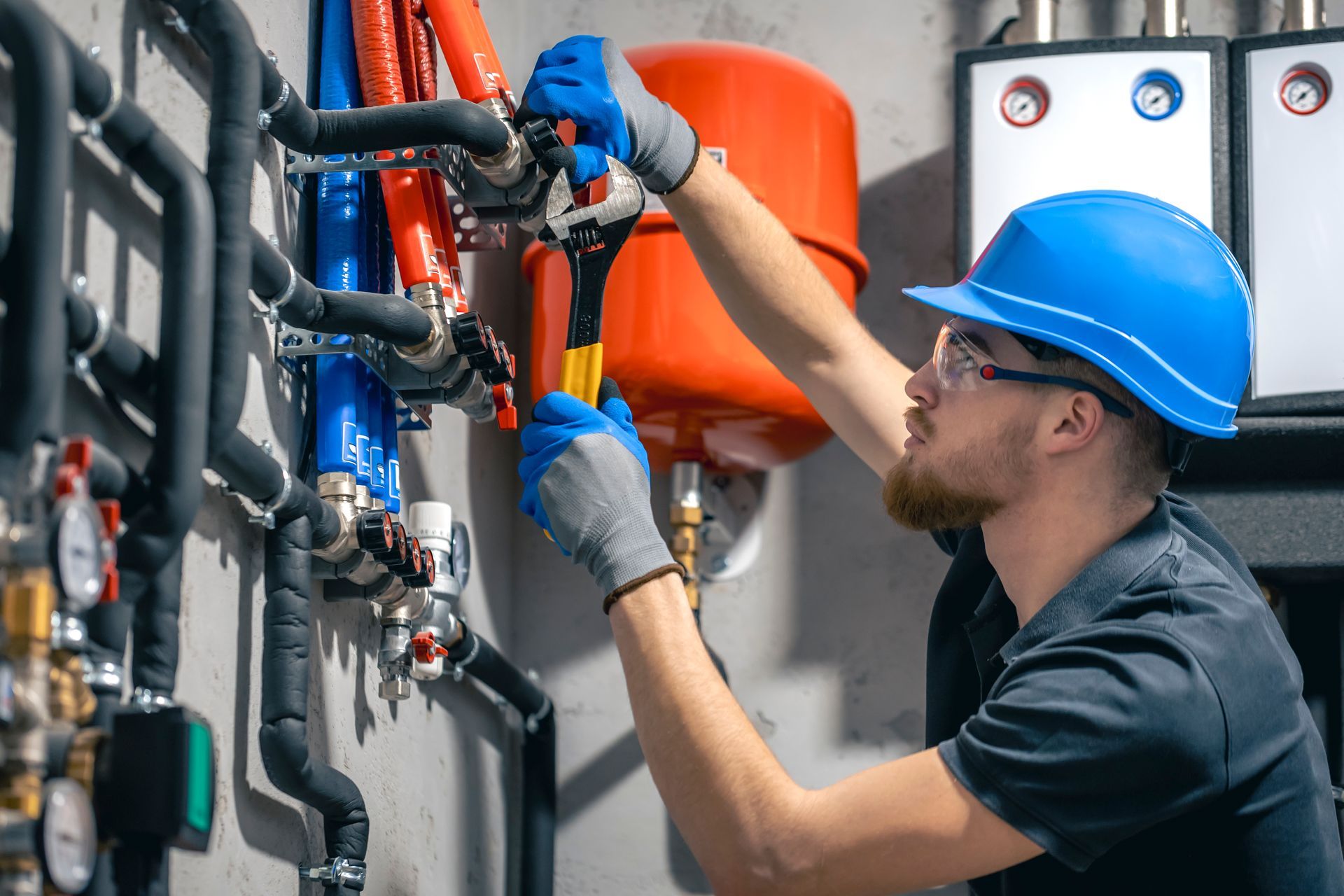 Plumber using a wrench on pipes. Wearing a blue hard hat and gloves, working in an equipment room.