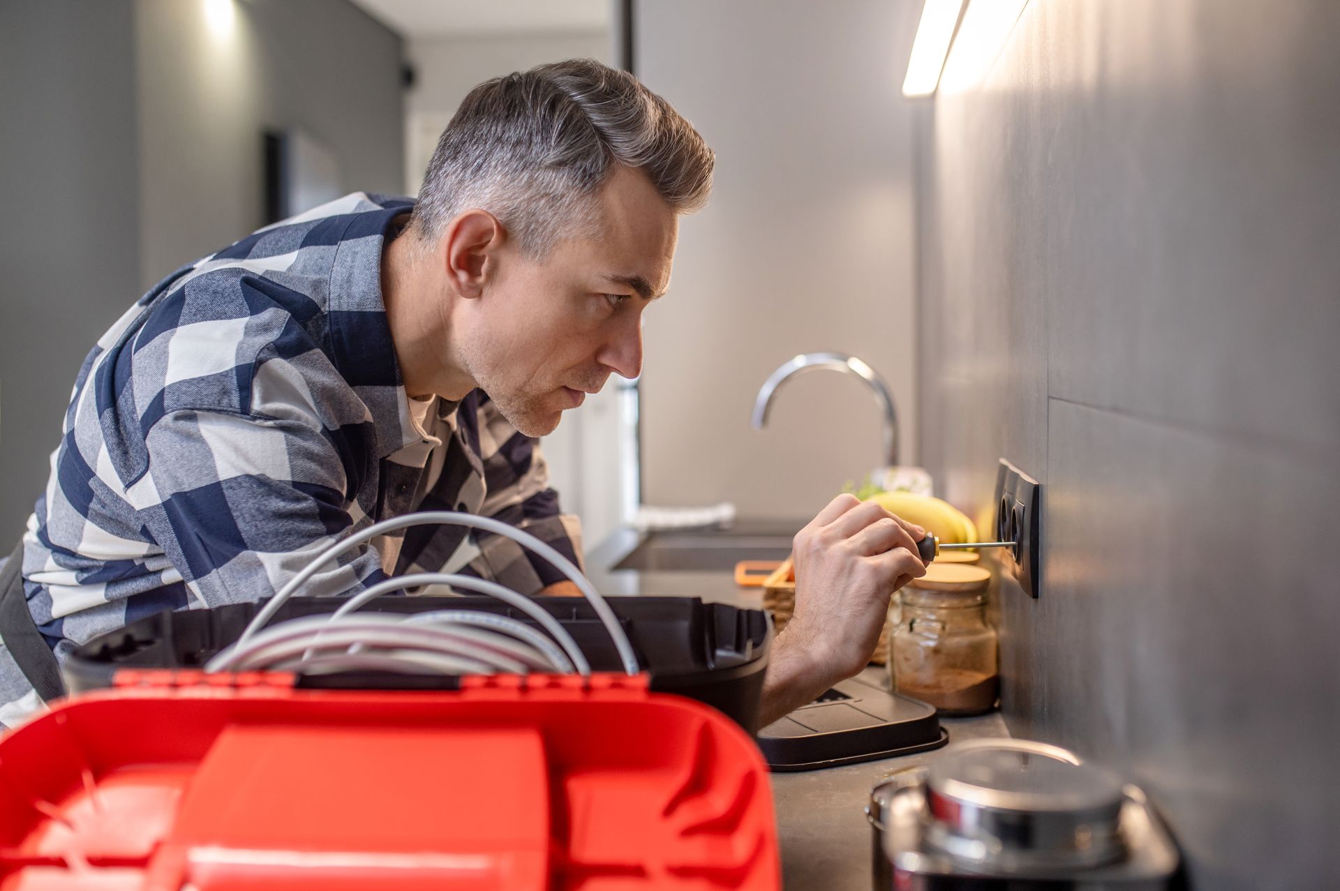 Man in plaid shirt working on an electrical outlet in a kitchen.