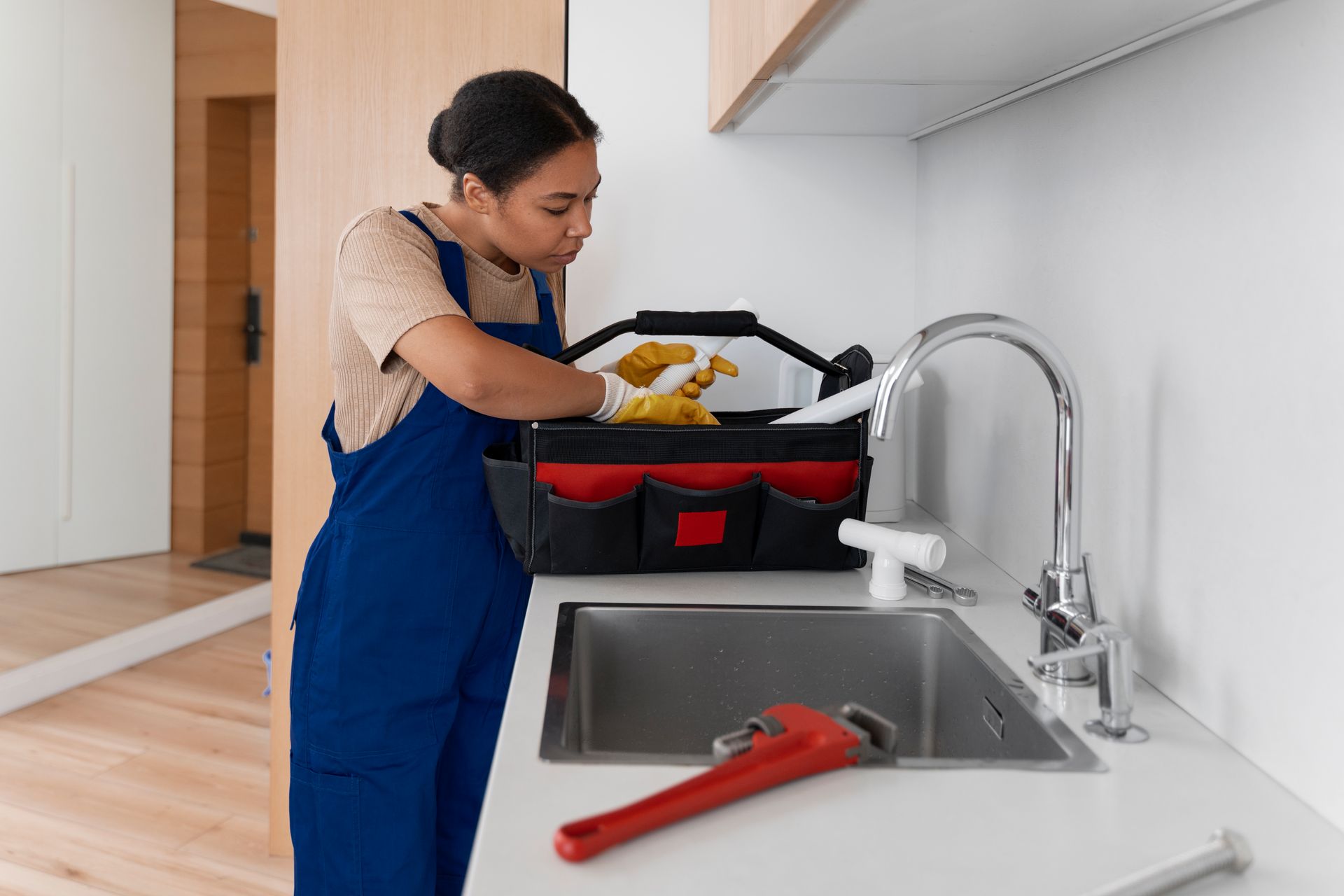 Plumber in blue overalls looking in toolbox next to a sink, red wrench on counter.