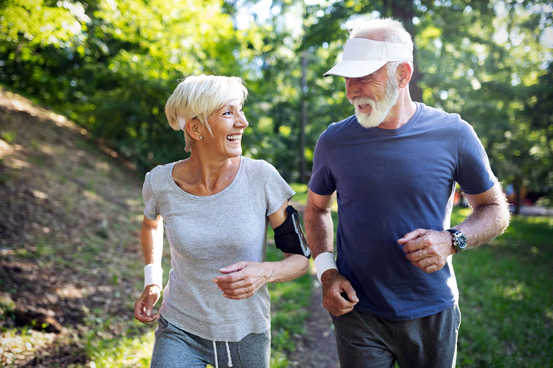Two people jogging on a sunlit trail in a green park, smiling and wearing athletic clothes.