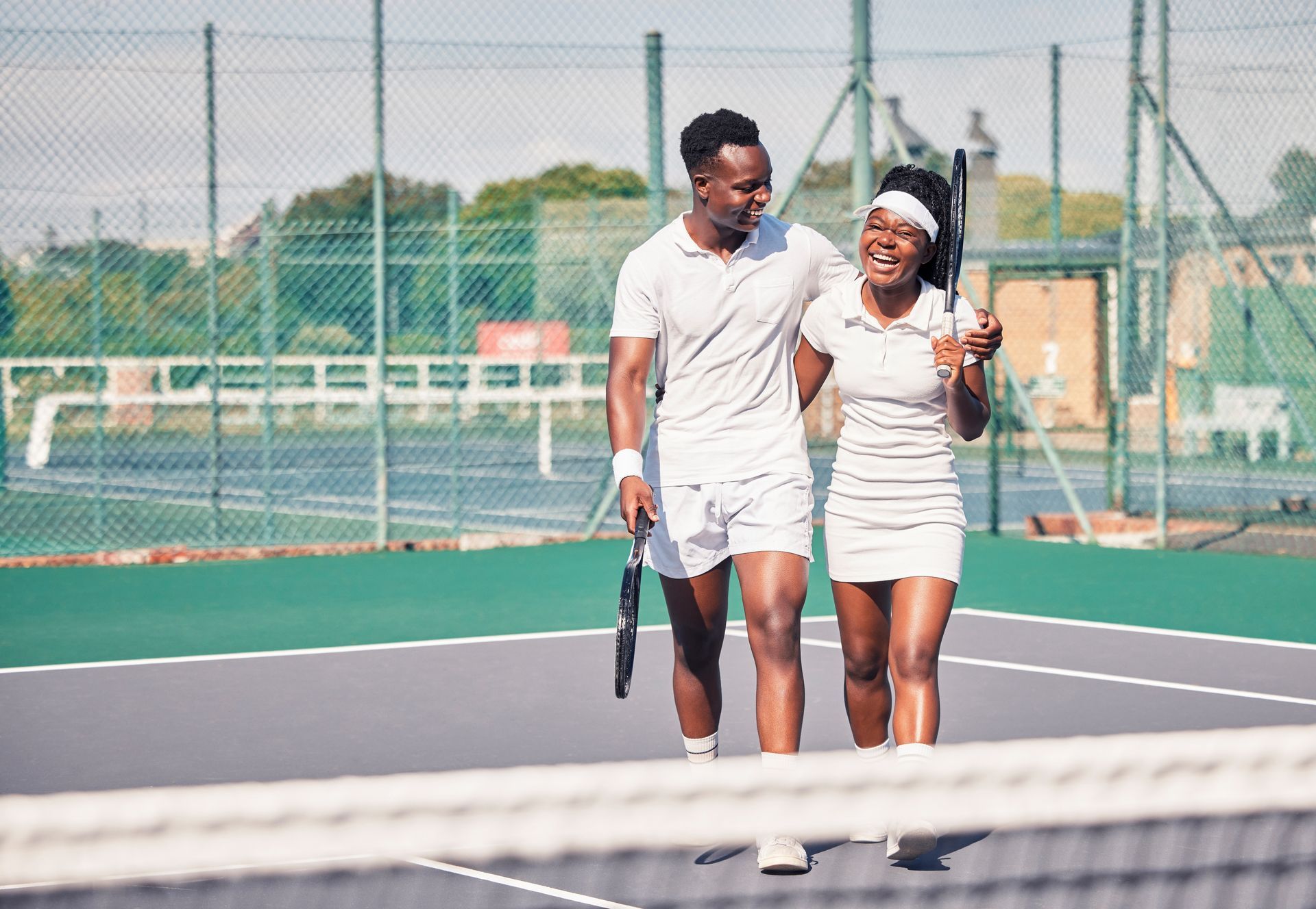 Two tennis players walking arm-in-arm on an outdoor court, smiling and holding rackets