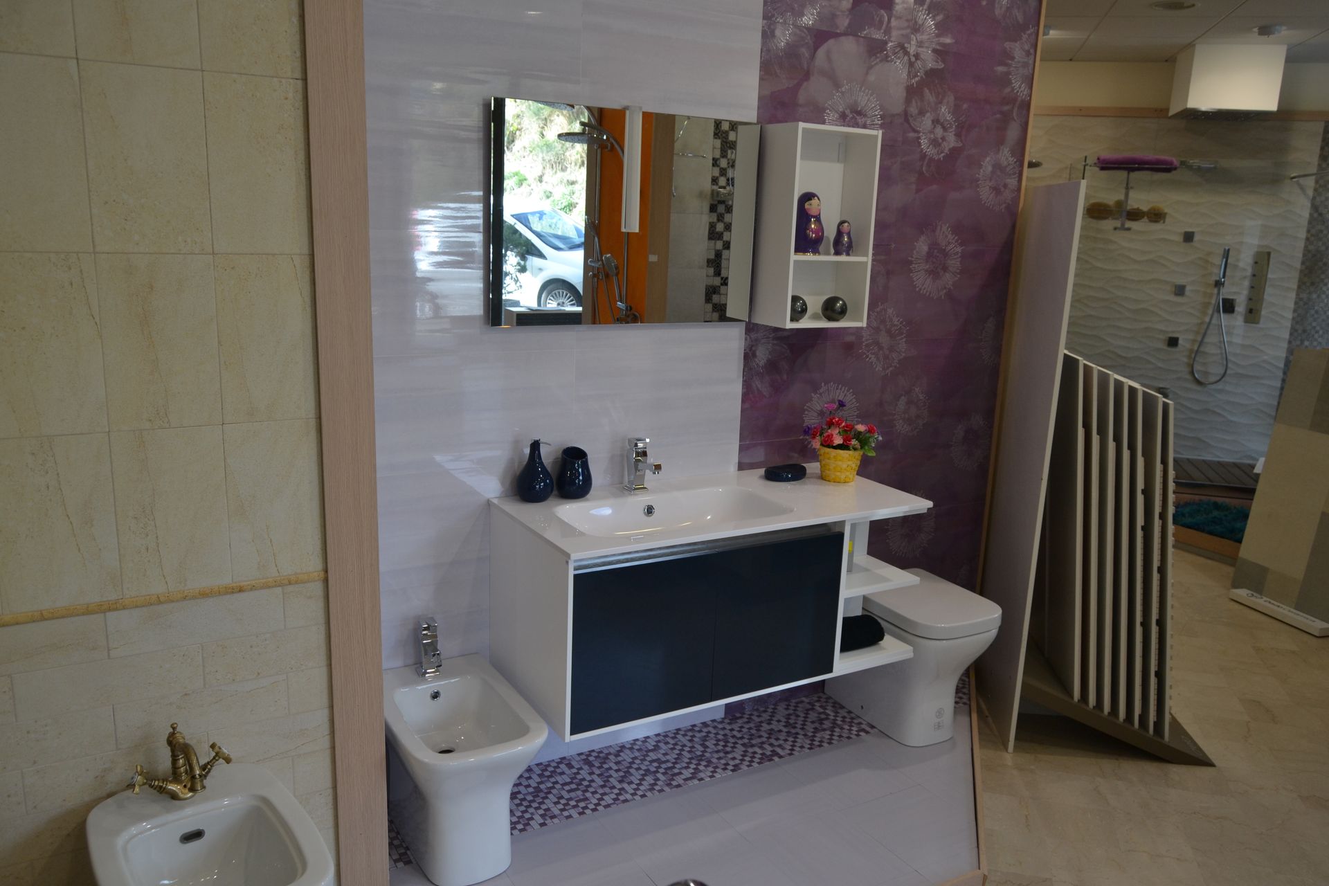 Bathroom display with white sink, black cabinet, bidet, toilet, mirror, and purple floral wallpaper.