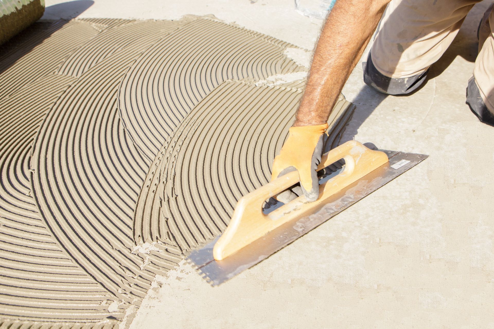Person using notched trowel to spread mortar for tiling on a concrete surface.