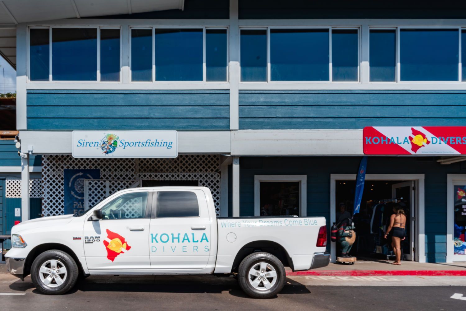 A Kohala Diver white truck is parked in front of a blue building.